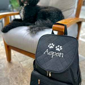 May include: Black quilted dog carrier backpack with a zippered pocket and a dispenser for dog waste bags. The backpack has white paw prints and the name "Aspen" embroidered on the front.