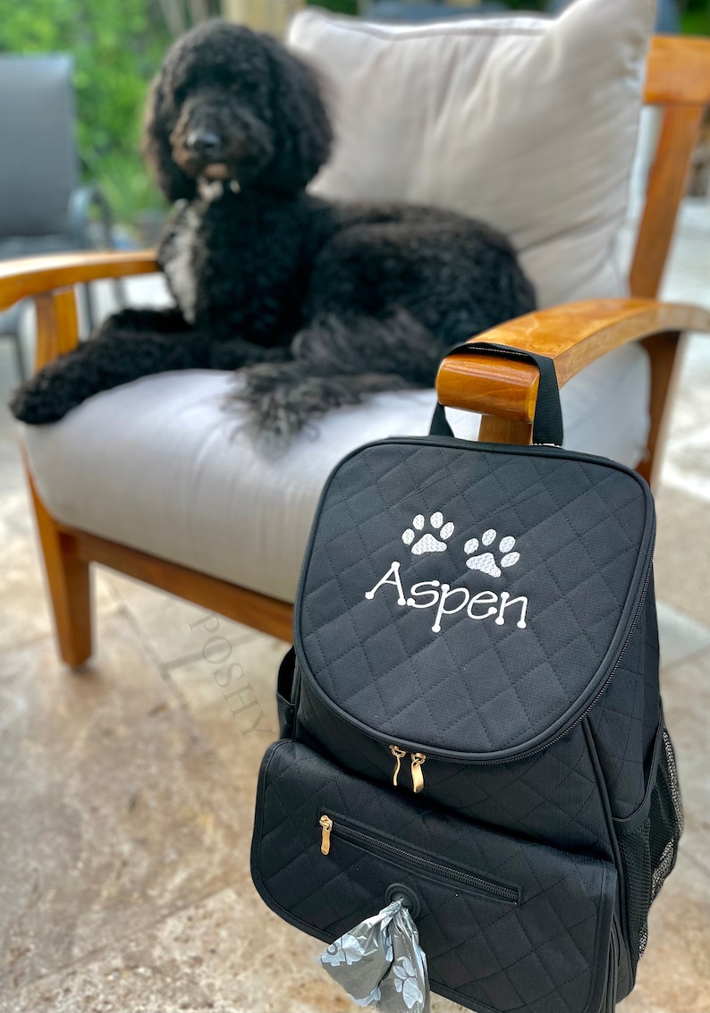 May include: Black quilted dog carrier backpack with a zippered pocket and a dispenser for dog waste bags. The backpack has white paw prints and the name "Aspen" embroidered on the front.
