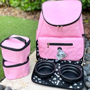 May include: Pink quilted dog carrier backpack with two collapsible food bowls and two pink storage containers. The backpack has a zippered pocket on the front and a mesh pocket on the side.