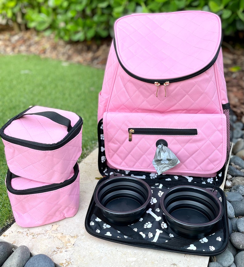 May include: Pink quilted dog carrier backpack with two collapsible food bowls and two pink storage containers. The backpack has a zippered pocket on the front and a mesh pocket on the side.