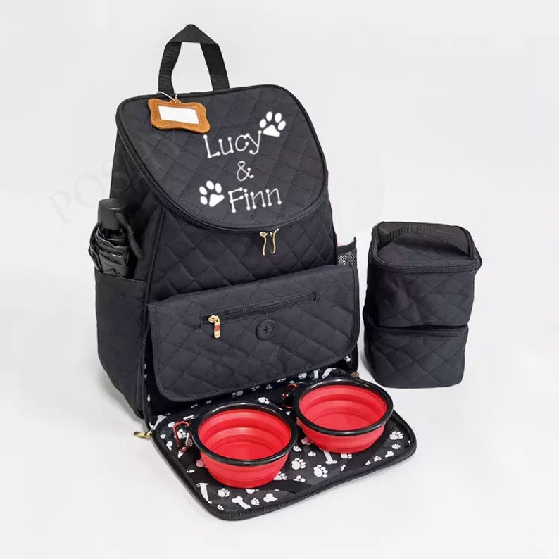 May include: Black quilted dog backpack with two red collapsible bowls, a side pocket, and a separate insulated food container. The backpack has a name tag and is personalized with the text "Lucy & Finn" in white.