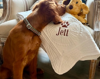 May include: A white quilted blanket featuring the embroidered name Jett in brown thread below a brown paw print icon. The blanket is draped over a chair next to a golden-brown plush dog toy and a reddish-brown dog wearing a green and white collar.