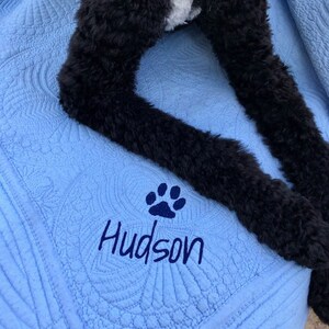 May include: A blue quilted blanket with a paw print and the name "Hudson" embroidered on it. A black dog is lying on the blanket.