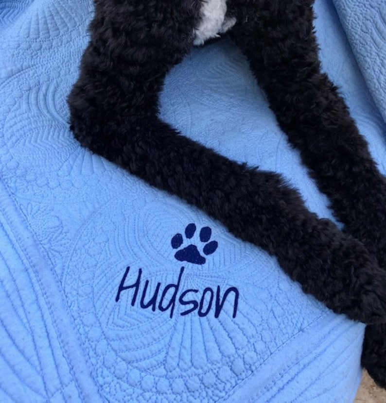 May include: A blue quilted blanket with a paw print and the name "Hudson" embroidered on it. A black dog is lying on the blanket.