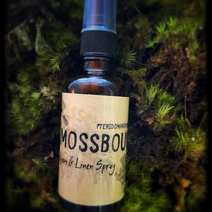 May include: A brown glass spray bottle with a black cap and a clear plastic top. The label reads "MOSSBOU Room & Linen Spray" with botanical illustrations. The bottle is set against a backdrop of green moss.