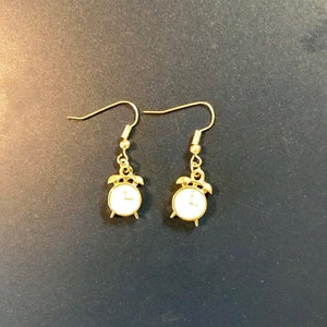 May include: Gold alarm clock earrings with white clock faces. The earrings have a simple hook design.