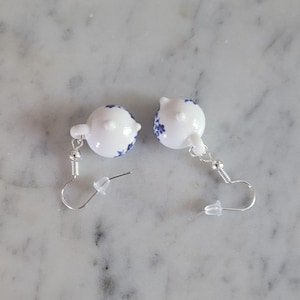 Op de afbeelding: Een paar witte keramische oorbellen met blauwe bloempatronen. Elke oorbel heeft een kleine tuit en een handvat en is bevestigd aan een zilverkleurige oorhaak. Transparante oorbelruggen zijn bevestigd.