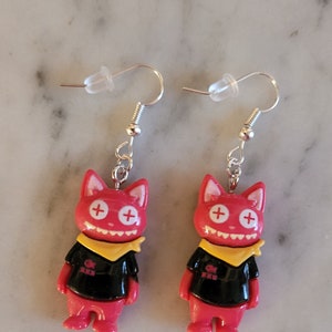May include: A pair of red cat-shaped earrings with black shirts, yellow bandanas, and white plus signs for eyes. The earrings are on a white background.