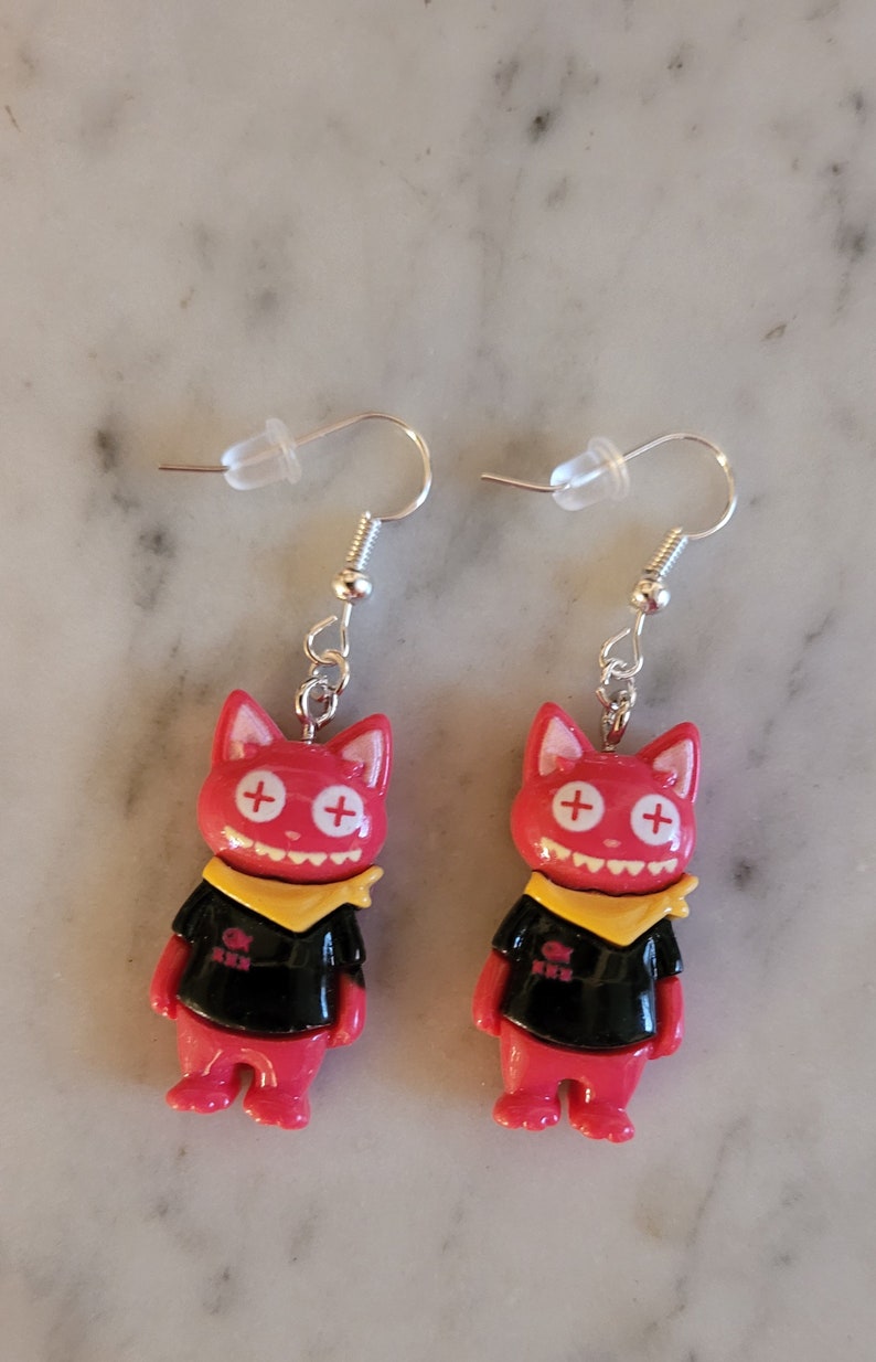 May include: A pair of red cat-shaped earrings with black shirts, yellow bandanas, and white plus signs for eyes. The earrings are on a white background.