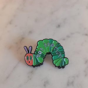 May include: A green and black enamel pin in the shape of a cartoon caterpillar. The caterpillar has a red face, large eyes, and the words "eat the rich" in white text across its body.