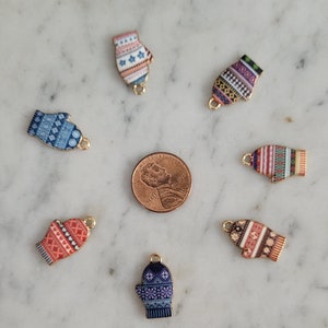 May include: A collection of colorful mitten charms with various patterns, including stars, stripes, and geometric designs. Each charm has a gold-colored loop for attaching to jewelry or other crafts. A penny is included for size reference.