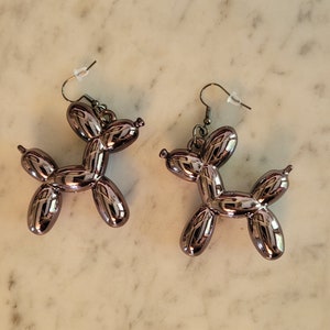 May include: A pair of metallic silver-colored dog-shaped earrings. The earrings are designed to resemble balloon animals, with a shiny, reflective surface. The earrings hang from silver-colored fish hook ear wires.
