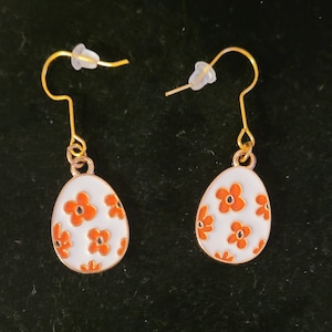 May include: A pair of gold earrings with white egg-shaped charms decorated with orange flowers.