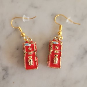 May include: A pair of gold dangle earrings with red fire truck charms. The trucks have white stars and blue details.