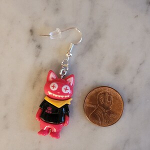 May include: Red and black cat-shaped dangle earrings with a yellow bandana and white plus signs for eyes. The earrings are shown next to a US penny for scale.
