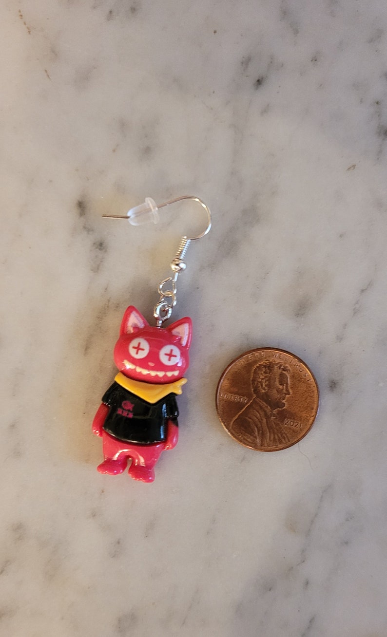 May include: Red and black cat-shaped dangle earrings with a yellow bandana and white plus signs for eyes. The earrings are shown next to a US penny for scale.