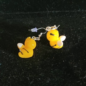 May include: A pair of yellow bee-shaped earrings with black stripes and white wings. The earrings have silver-colored hooks and a clear plastic backing. The bees are facing opposite directions.
