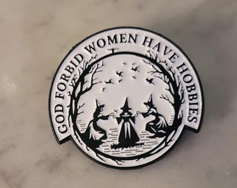 May include: A black and white enamel pin with the text "GOD FORBID WOMEN HAVE HOBBIES" encircling a central image. The image depicts three witches in a circle, surrounded by trees and bats.