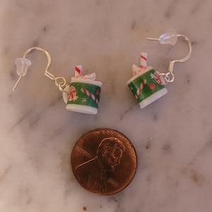 May include: A pair of silver dangle earrings with a miniature Christmas-themed hot chocolate mug charm. The mug is green and white with red and white candy cane accents. The mug is topped with whipped cream and a red and white candy cane.