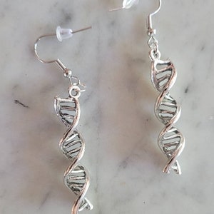 May include: A pair of silver-colored DNA helix earrings. Each earring features a detailed double helix design, with a hook for easy wear. The earrings are displayed on a marble surface.