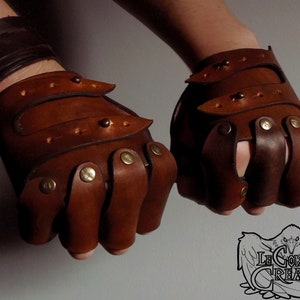 Könnte beinhalten: Ein Paar braune fingerlose Lederhandschuhe mit Messingakzenten. Die Handschuhe haben mehrere Riemen und Schnallen.