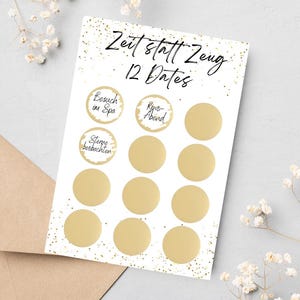 Puede incluir: Una tarjeta blanca con círculos dorados para rascar y el texto "Zeit statt Zeug 12 Dates" en escritura negra. La tarjeta está sobre una superficie gris claro con un sobre marrón y flores blancas.