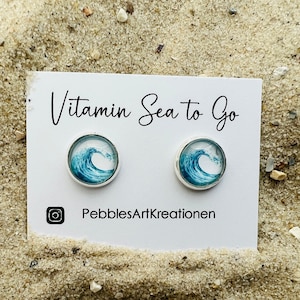 Puede incluir: Pendientes de botón redondos con un diseño de ola azul y blanca. Los pendientes están engastados en metal plateado y se muestran en una tarjeta blanca con el texto "Vitamin Sea to Go" y el identificador de Instagram "PebblesArtKreationen".