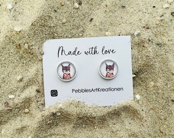 May include: A pair of earrings featuring a close-up image of a lynx with a red and white background. The earrings are made with glass cabochons and are set in silver-toned settings. The earrings are displayed on a white card with the text "Made with love" and "PebblesArtKreationen".