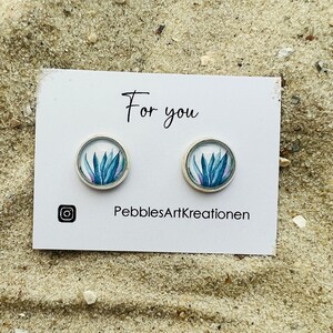 Könnte beinhalten: Silberfarbene Ohrstecker mit einem Aquarelldesign aus blauen und violetten Pflanzenblättern. Die Ohrringe werden auf einer weißen Karte mit dem Text "For you" und dem Instagram-Logo präsentiert. Die Karte enthält auch den Text "PebblesArtKreationen".