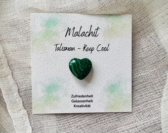 May include: A heart-shaped malachite stone on a card with the word "Malachit" and the phrase "Talisman - Keep Cool". The stone is green with dark green swirling patterns. The card also includes the words "Zufriedenheit, Gelassenheit, and Kreativität."