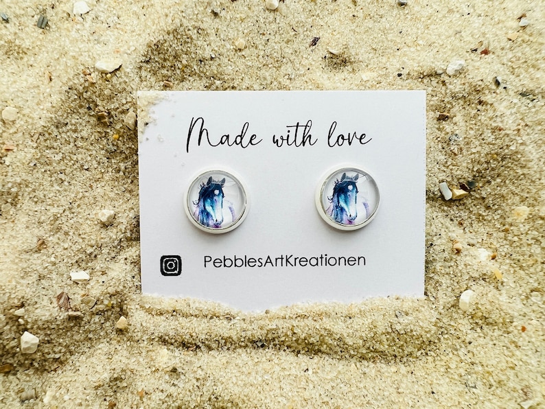 May include: A pair of silver stud earrings with a watercolor painting of a horse's head on each. The earrings are on a white card with the text "Made with love" and "PebblesArtKreationen".