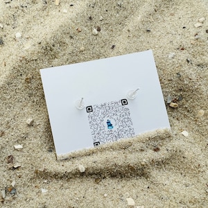 K&ouml;nnte beinhalten: Eine wei&szlig;e Karte mit einem QR-Code und zwei kleinen silbernen Ohrringen darauf. Die Karte liegt auf einer sandigen Oberfl&auml;che.