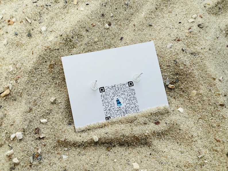 K&ouml;nnte beinhalten: Eine wei&szlig;e Karte mit einem QR-Code und zwei kleinen silbernen Ohrringen darauf. Die Karte liegt auf einer sandigen Oberfl&auml;che.