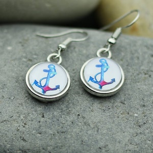 May include: A pair of silver-toned dangle earrings. Each earring features a round, white background with a blue and pink anchor design. The earrings are displayed on a gray stone surface.