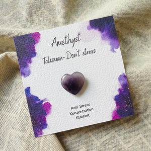 Könnte beinhalten: Ein herzförmiger Amethyststein liegt auf einer Karte mit den Worten "Amethyst Talisman-Don't stress". Die Karte hat lila Aquarellakzente und die Worte "Anti-Stress, Konzentration, Klarheit".