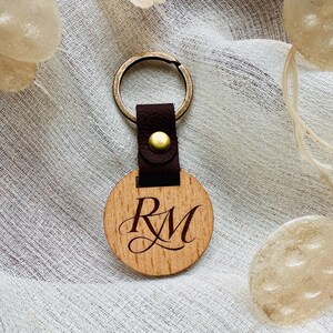 May include: A wooden round keychain with the engraved initials "RM". The keychain features a dark brown leather strap and a brass-colored metal ring. The wood has a natural grain, and the initials are in an elegant script.