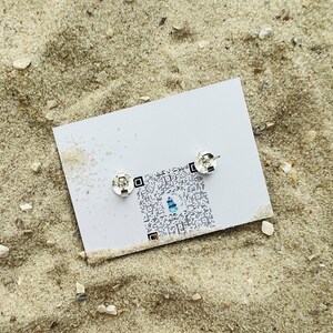 K&ouml;nnte beinhalten: Ein Paar silberne Ohrstecker auf einer wei&szlig;en Karte mit einem QR-Code. Die Ohrstecker sind klein und rund.