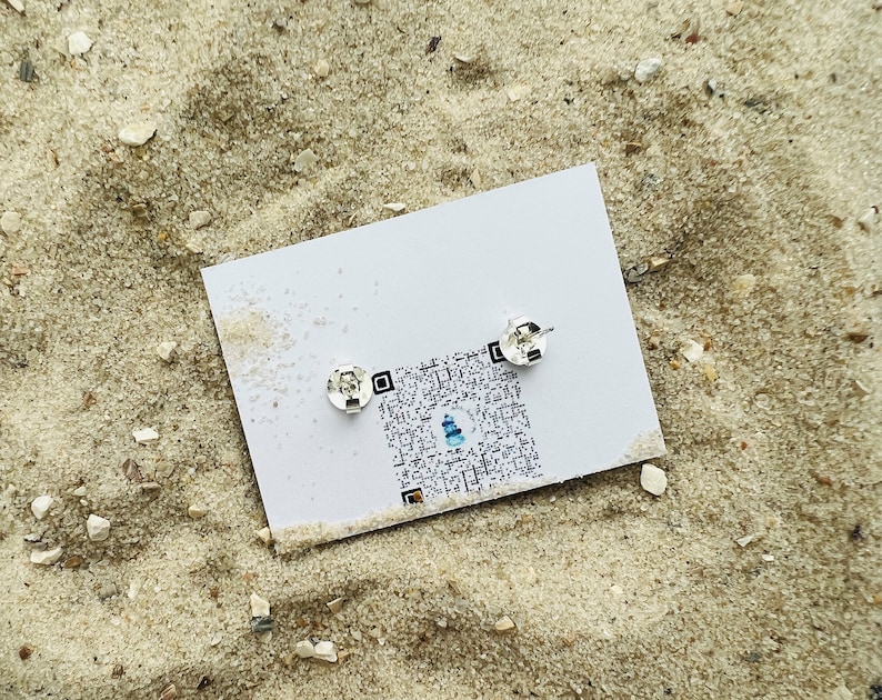 K&ouml;nnte beinhalten: Ein Paar silberne Ohrstecker auf einer wei&szlig;en Karte mit einem QR-Code. Die Ohrstecker sind klein und rund.
