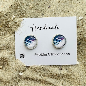 Könnte beinhalten: Zwei runde Ohrstecker mit silbernem Rand. Die Ohrringe haben ein farbenfrohes abstraktes Design mit blauen, weißen und lila Pinselstrichen. Sie liegen auf einer weißen Karte mit der Aufschrift "Handmade" und "PebblesArtKreationen".