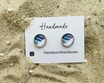 May include: Two round stud earrings with a silver border. The earrings feature a colorful abstract design with blue, white, and purple brushstrokes. The earrings are displayed on a white card that says "Handmade" and "PebblesArtKreationen".
