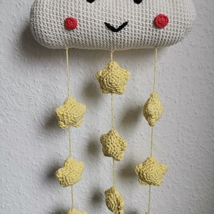 Könnte beinhalten: Gehäkelte Wanddekoration in Form einer weißen Wolke mit einem lächelnden Gesicht, schwarzen Augen und roten Wangen. Gelbe Sterne und Tropfen hängen an der Wolke. Ideal für das Kinderzimmer.