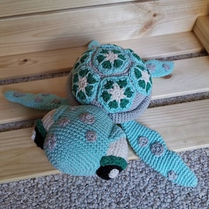 Könnte beinhalten: Ein gehäkeltes Meeresschildkrötenspielzeug mit einer grün-weißen Schale und einem hellblauen Körper. Die Schildkröte hat schwarze und weiße Räder an der Unterseite.