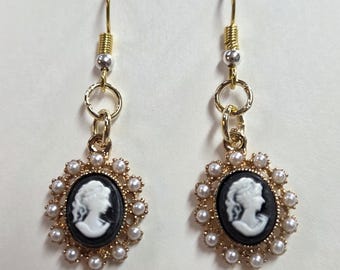 May include: A pair of gold-toned drop earrings. Each earring features a black cameo pendant with a white profile, surrounded by faux pearls. The earrings have gold-colored hooks and decorative accents.
