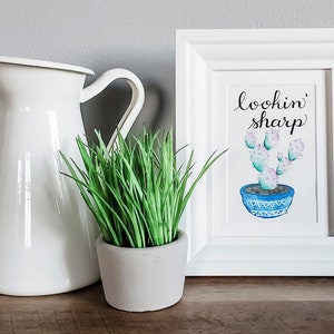 Può includere: Una stampa con cornice bianca con le parole "lookin' sharp" sopra un cactus ad acquerello in un vaso blu. Una brocca bianca e una piccola pianta in vaso con erba verde sono accanto alla stampa.