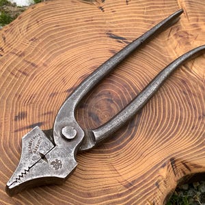 Könnte beinhalten: Antike Metallzange mit langen Griffen und gezahnten Backen. Die Zange ist mit Text und einem Symbol auf dem Kopf markiert. Das Werkzeug wird auf einer Holzoberfläche mit natürlichem Maserungsmuster dargestellt.