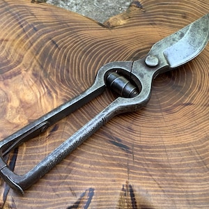 May include: A pair of vintage metal pruning shears with a weathered, dark gray finish. The tool features a curved, sharp blade, a central pivot bolt, and a textured handle grip. The design includes a metal loop at the base of the handles.