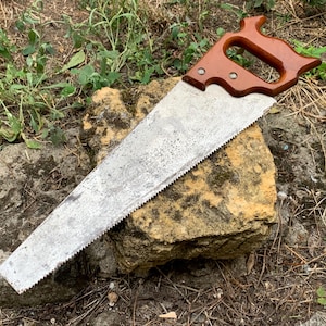 May include: A hand saw featuring a weathered, metallic triangular blade with a serrated edge and a polished, dark brown wooden handle secured by two metal rivets. The tool exhibits signs of age and use, with a textured, oxidized surface finish.