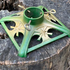 Könnte beinhalten: Ein quadratischer Weihnachtsbaumständer aus grünem Metall mit einer zentralen zylindrischen Halterung, drei verstellbaren Metall-Ringschrauben und sternförmigen Ausschnitten an allen vier Seiten, veredelt mit einem dezenten goldenen Sprühfinish.