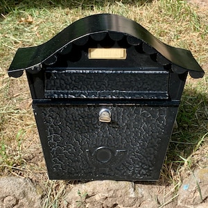Könnte beinhalten: Ein schwarzer Briefkasten aus Metall mit strukturierter Oberfläche und einem geschwungenen Dach. Die Vorderseite verfügt über einen horizontalen Einwurfschlitz, ein zentrales Schloss mit eingestecktem silbernen Schlüssel und ein Namensschild.