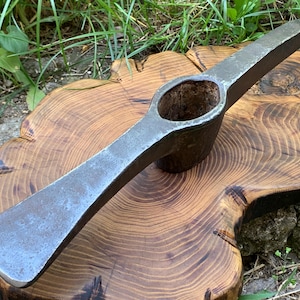 May include: A heavy-duty, forged steel pickaxe head featuring a pointed end and a wide, flat chisel blade on opposite sides. The tool has a central, oval-shaped eye for a handle attachment, showcasing a weathered, dark grey metallic finish.